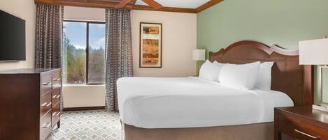 A hotel room with a bed, a desk, a ceiling fan, a window with curtains, and a painting on the wall.