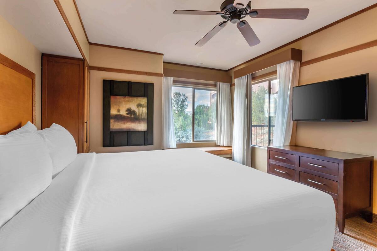 Um quarto de hotel espaçoso, com uma cama grande, ventoinha no teto, televisão de tela plana e janela com cortinas.