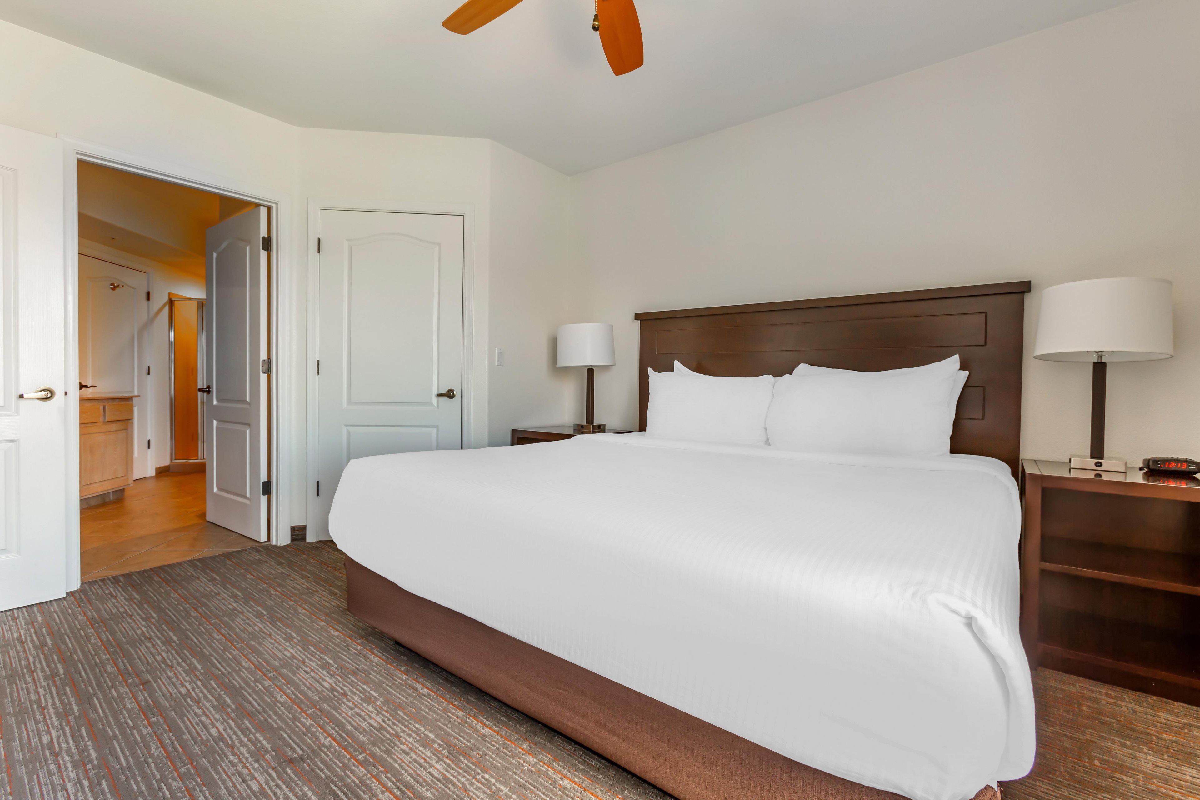 A hotel room with a large bed, two bedside tables with lamps, a wooden headboard, and a door leading to a hallway.