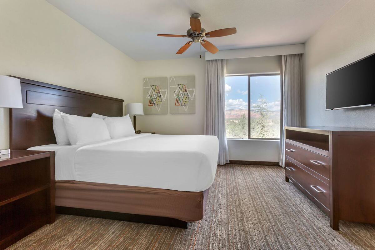 Habitación de hotel con cama, escritorio, aparador, ventilador de techo y una ventana grande con vistas.