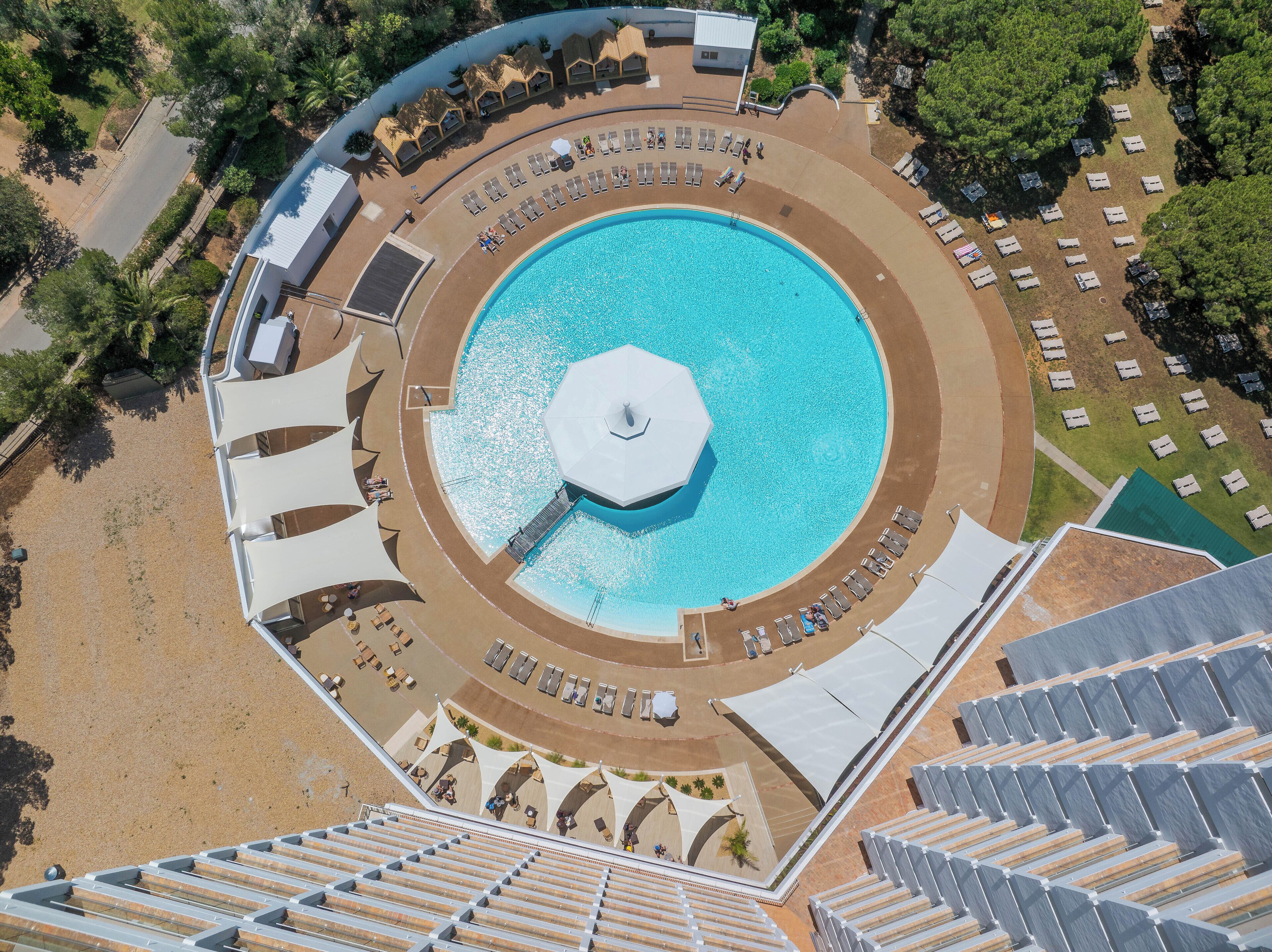 An aerial view of a resort with a central pool, sun loungers, and beach umbrellas.