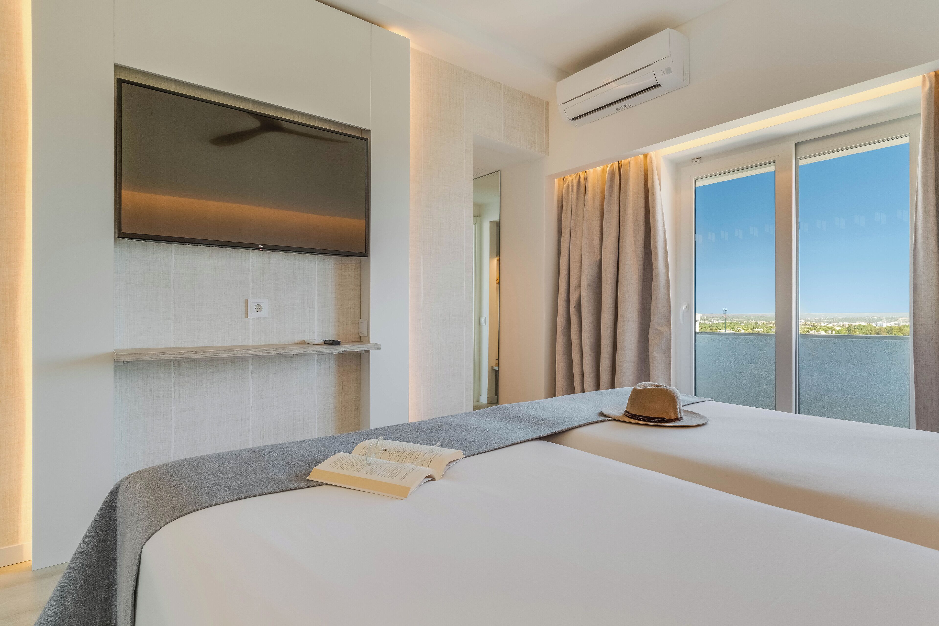 Habitación de hotel con cama, televisor de pantalla plana montado en la pared, aire acondicionado y vista al exterior a través de una puerta corrediza de vidrio.