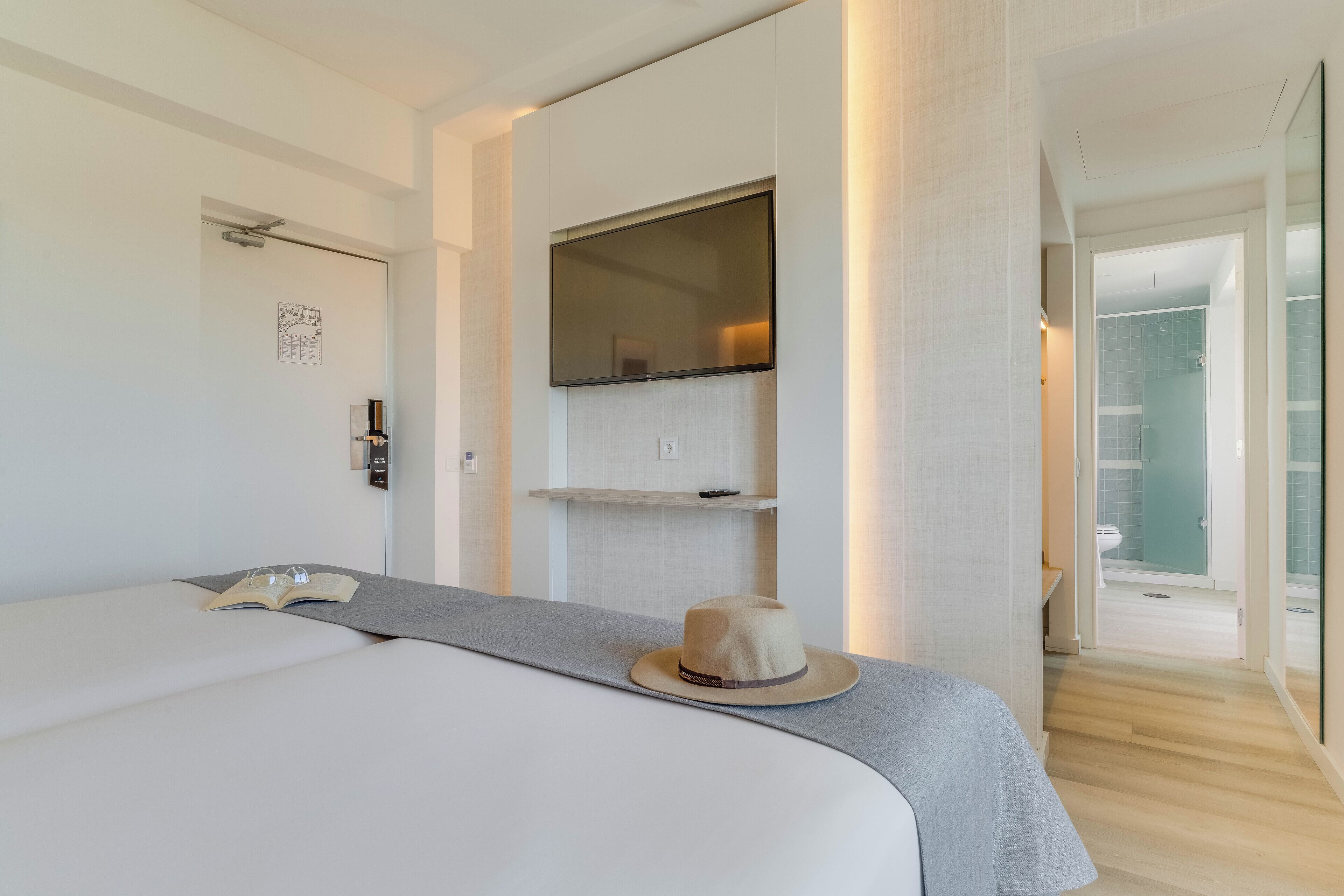 Una habitación de hotel moderna con una cama grande, un televisor de pantalla plana montado en la pared, un estante pequeño y una puerta que da a un baño.