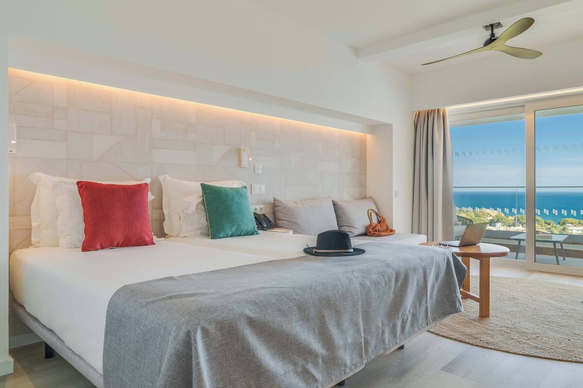 A modern hotel room with a large bed, a laptop on a small table, and a view of the ocean through large windows.