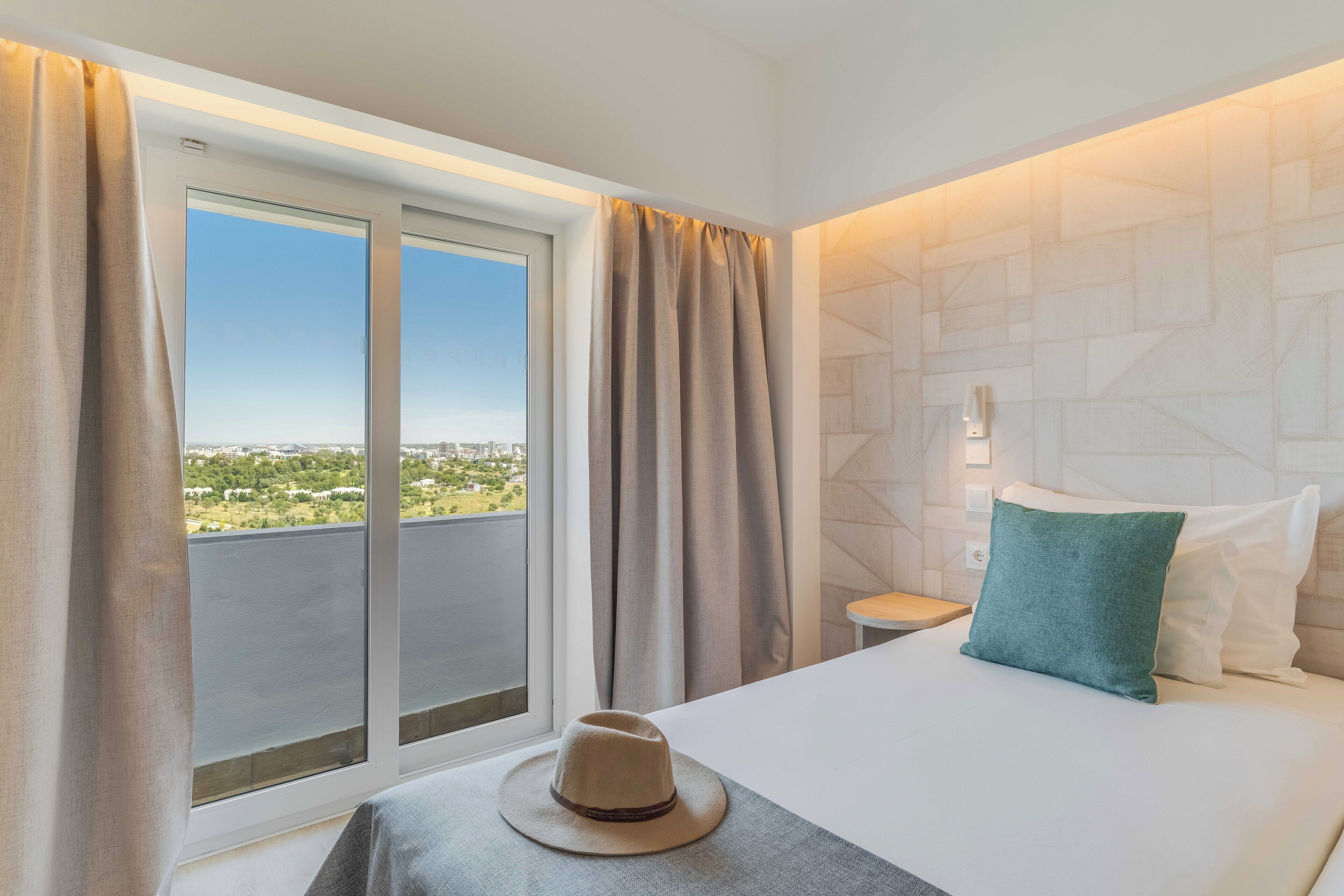 A hotel room with a bed, a nightstand, a lamp, a window with curtains, and a view of the city.