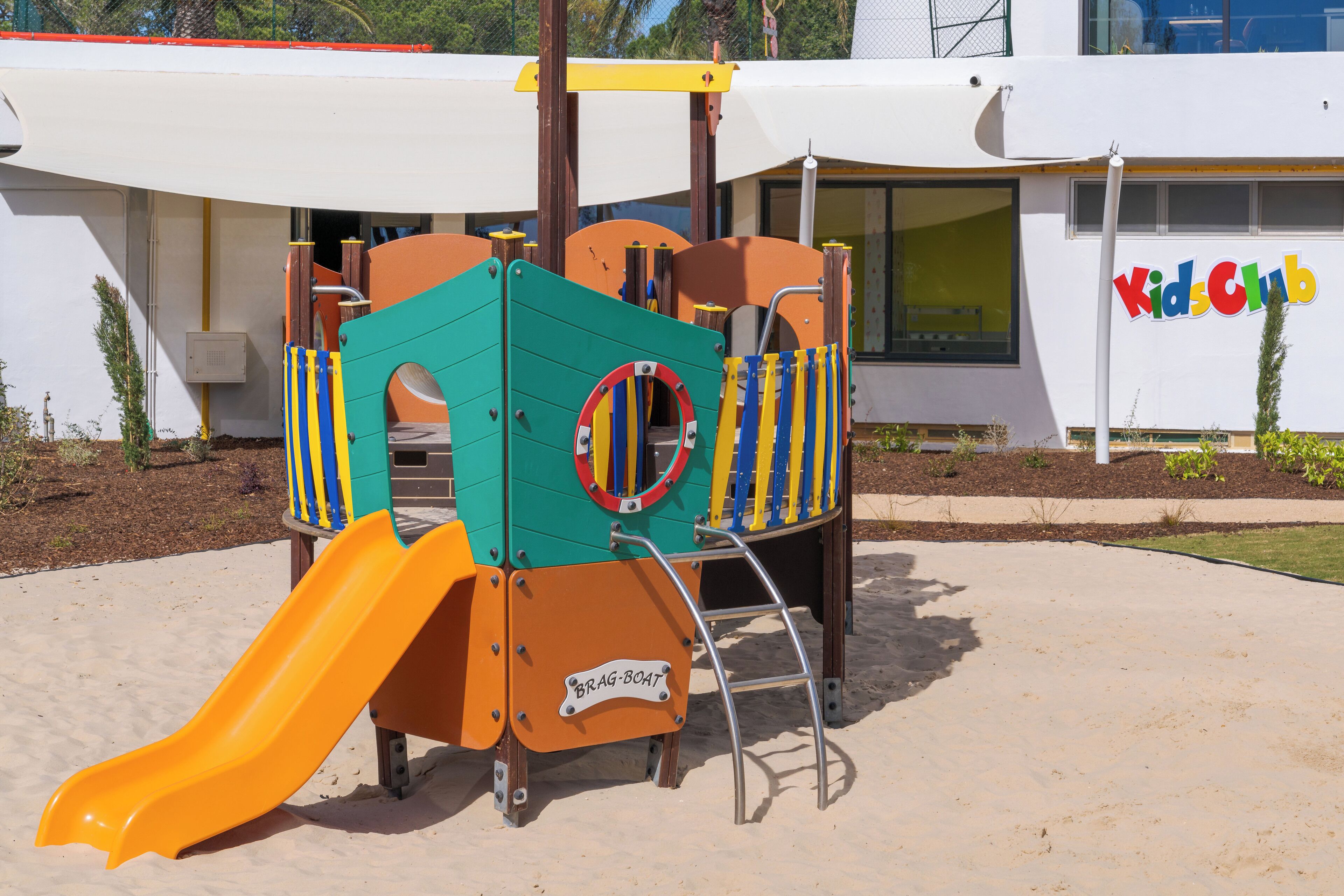 En legeplads med rutsjebane og klatrestrukturer, foran en bygning med skiltet 'Kids Club'.