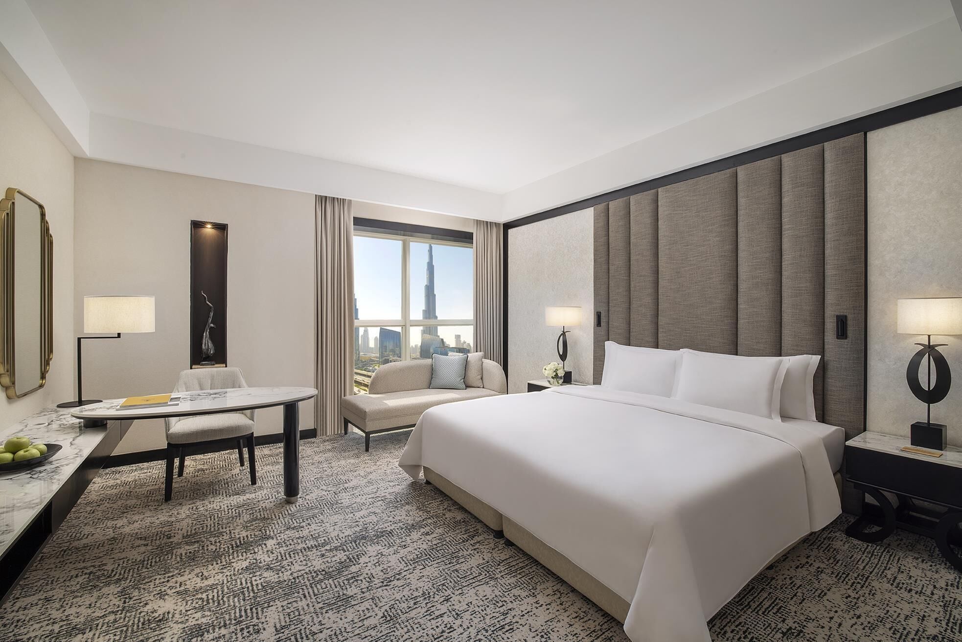 A hotel room with a large bed, a desk with a chair, a small table, and a view of the cityscape.