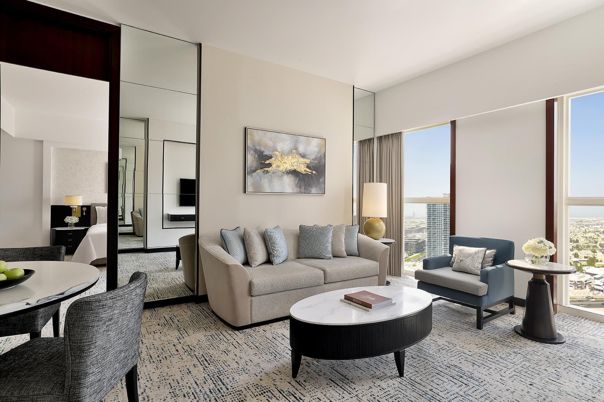 A modern hotel room with a sofa, armchair, dining table, and a large window offering a city view.