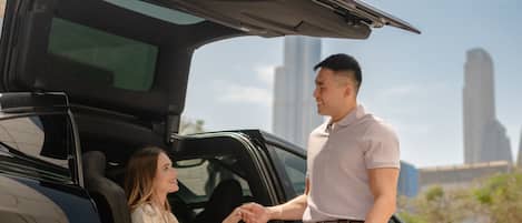 Un hombre y una mujer de pie junto a un coche descapotable negro con el maletero abierto, en un entorno urbano con edificios altos al fondo.