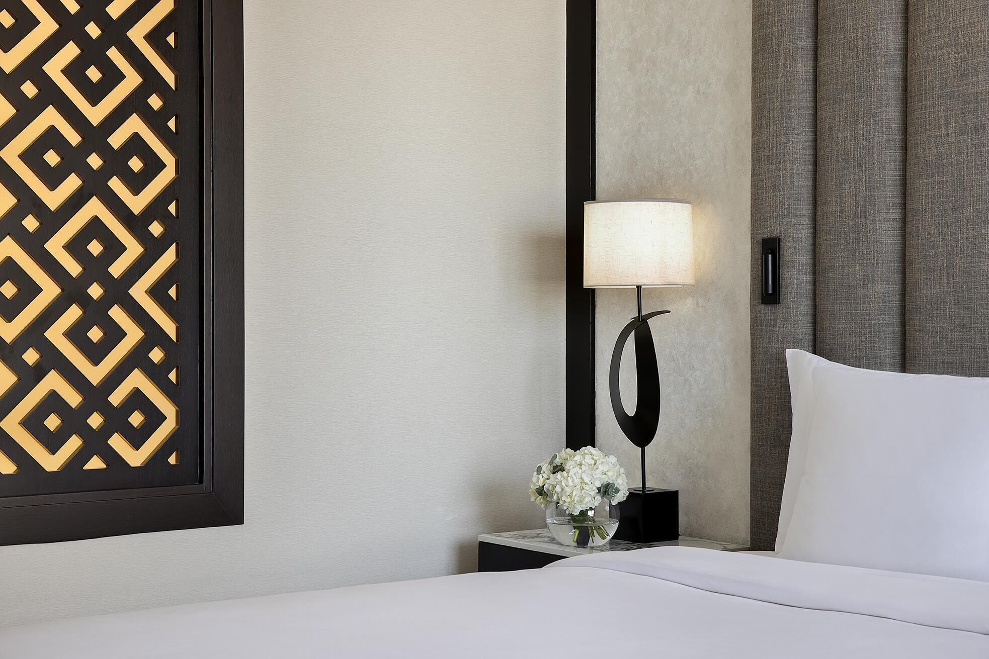 A hotel room with a bed, a nightstand with a lamp, a vase with flowers, and a decorative wall panel.