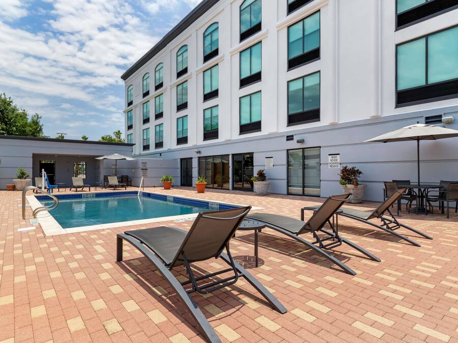 Zona de piscina con sillones de descanso y sombrillas, un patio de ladrillo y un edificio con ventanas amplias y un letrero.