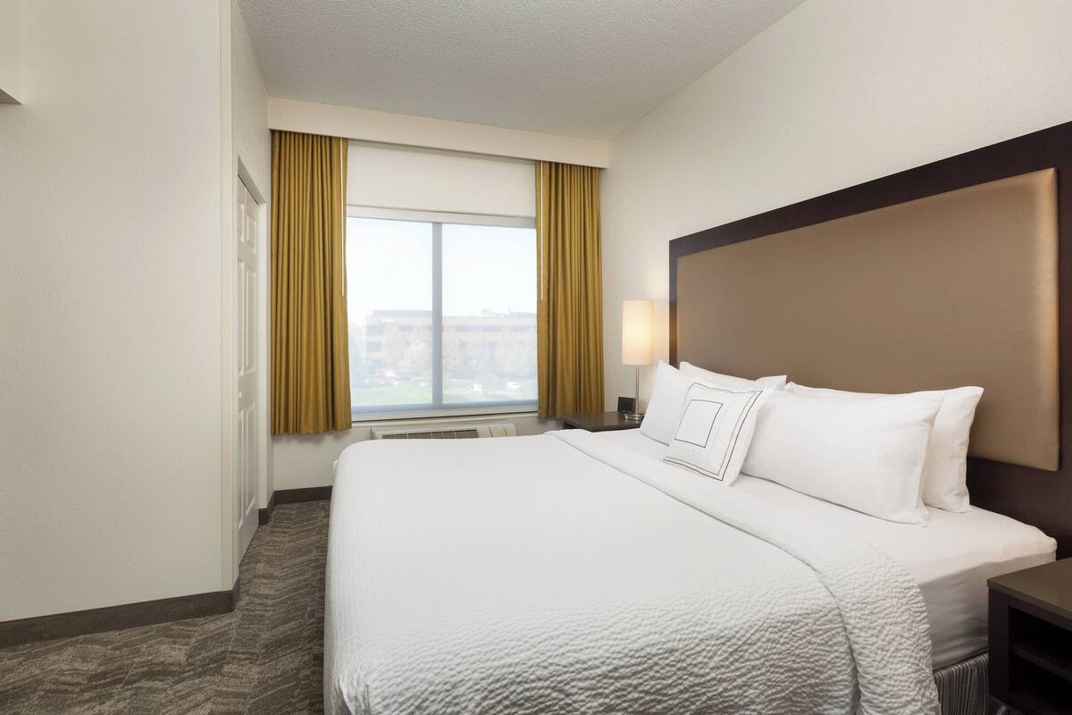 Ein Hotelzimmer mit einem großen Bett, einem Kopfteil, einem Nachttisch, einer Lampe und einem Fenster mit Vorhängen.