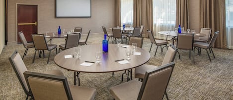 Un salón de conferencias con mesas y sillas redondas, una pantalla de proyector y un amplio ventanal con cortinas.