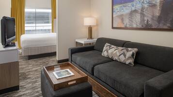Quarto de hotel com uma cama, um sofá, uma televisão, um abajur e um quadro na parede.