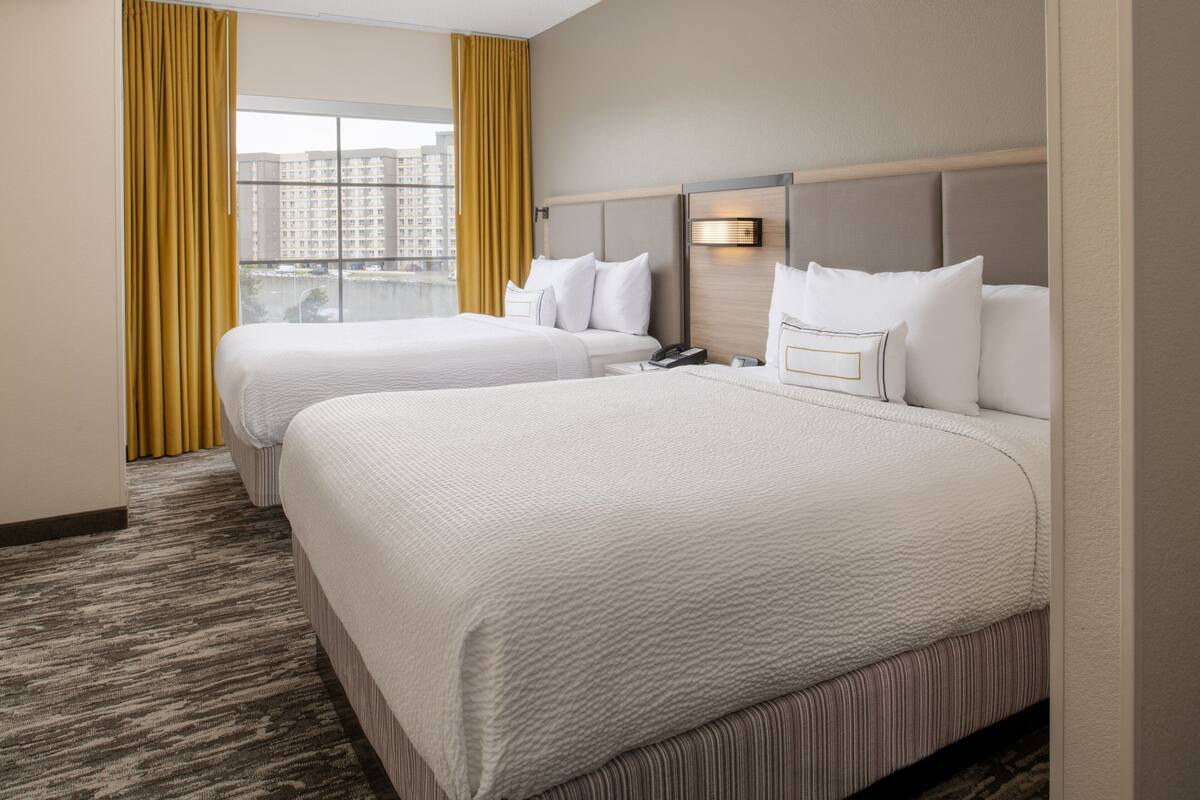 A hotel room with two beds, a large window with curtains, a headboard with a light, and a carpeted floor.
