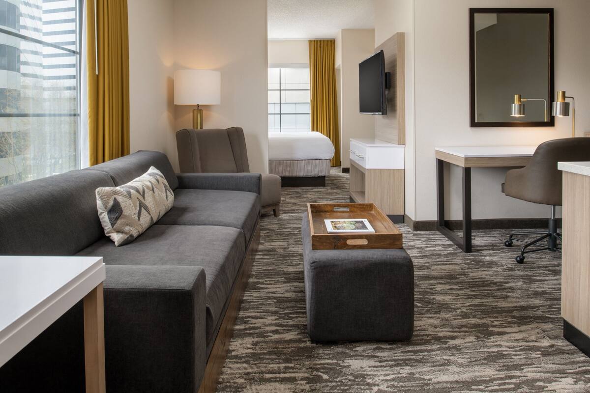 A hotel room with a grey sofa, a small ottoman, a desk, a bed, and a TV.