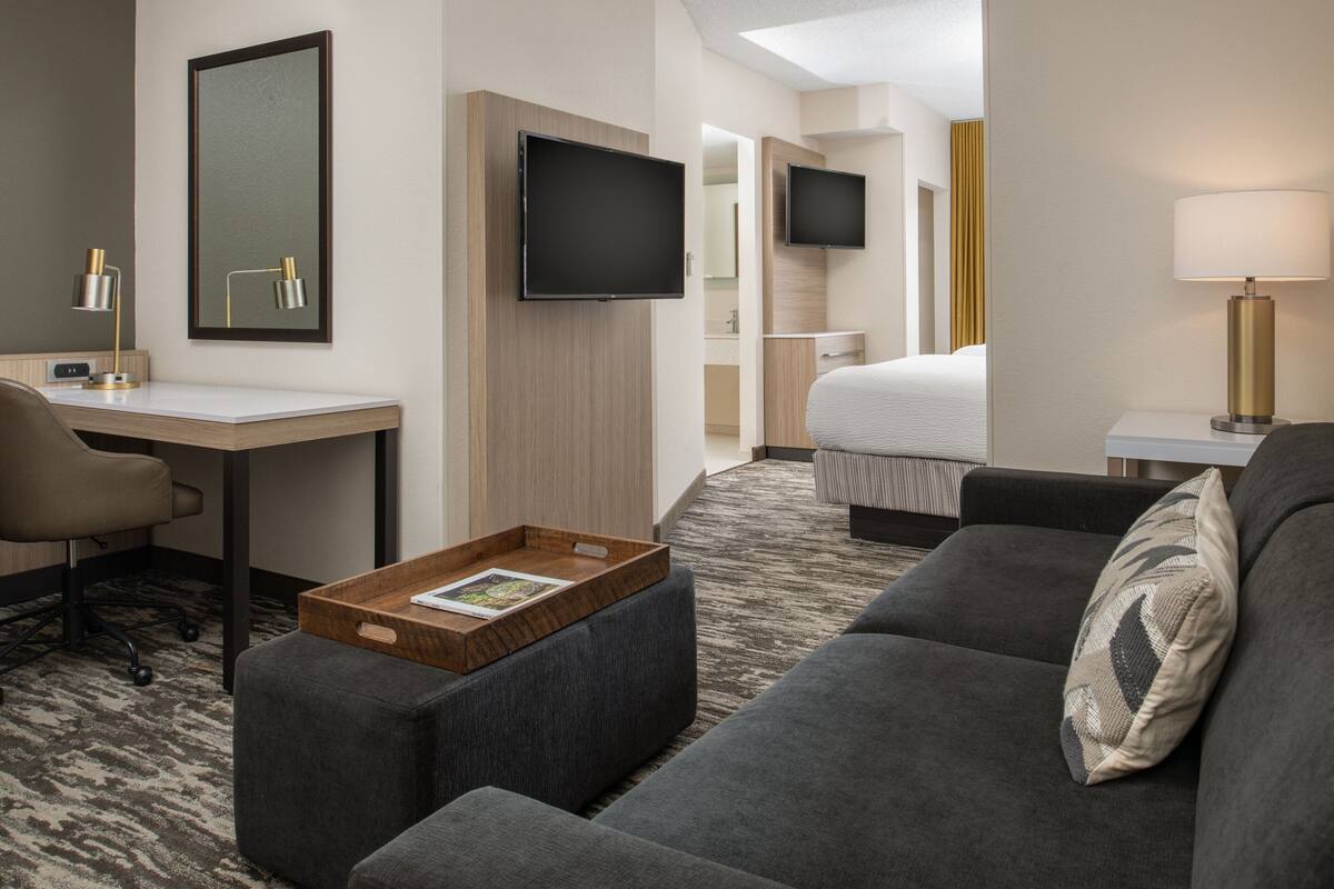 A hotel room with a grey sofa, a wooden side table with a tray, a desk with a chair, a flat-screen TV mounted on the wall, and a bed with a white duvet.