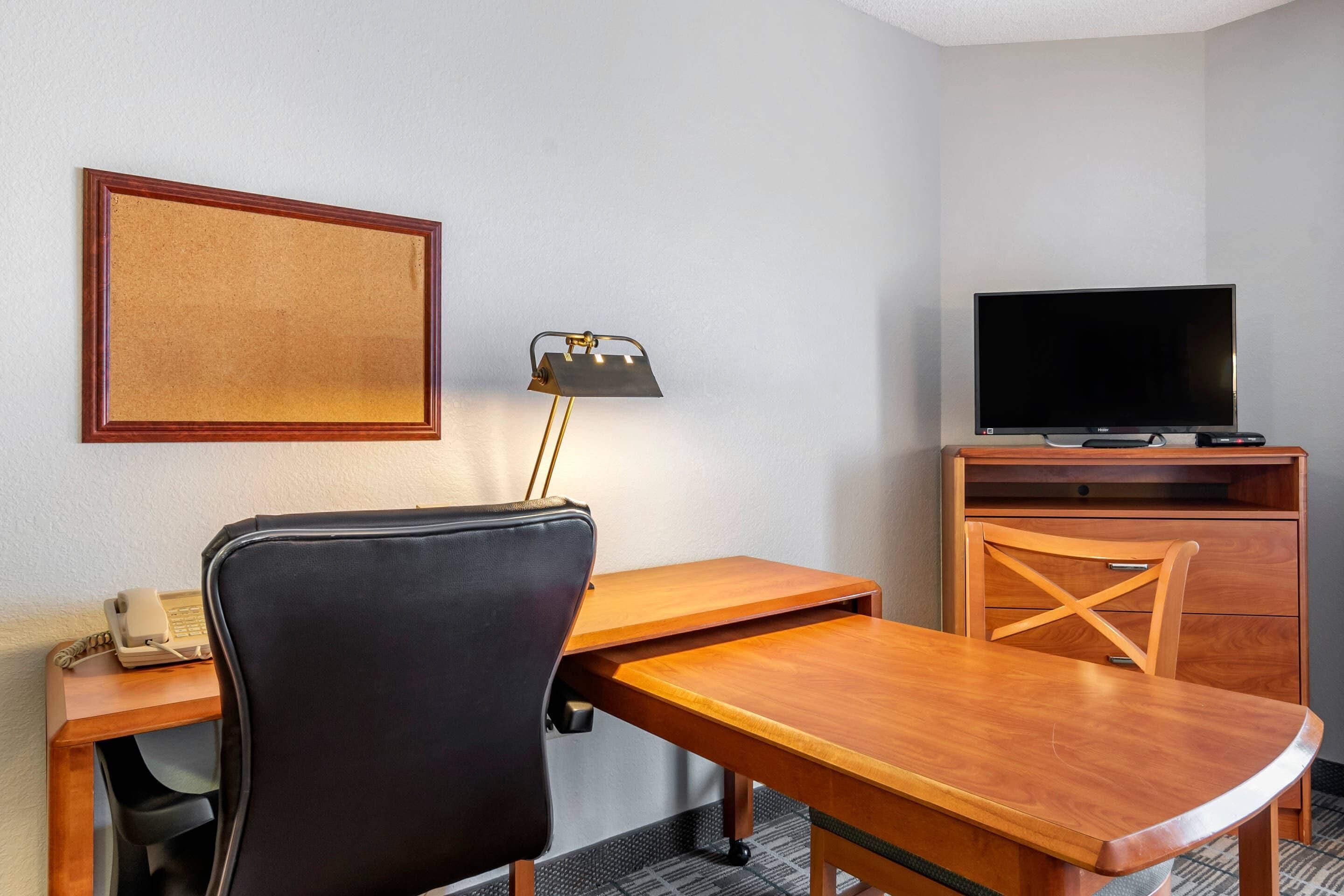 Una camera d'albergo con una scrivania in legno, una sedia da ufficio nera, un televisore su un mobile in legno e un quadro appeso al muro.