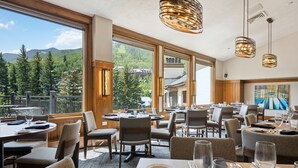 A restaurant with tables set for dining, large windows offering a mountain view, and hanging pendant lights. - The Charter at Beaver Creek (Avon)