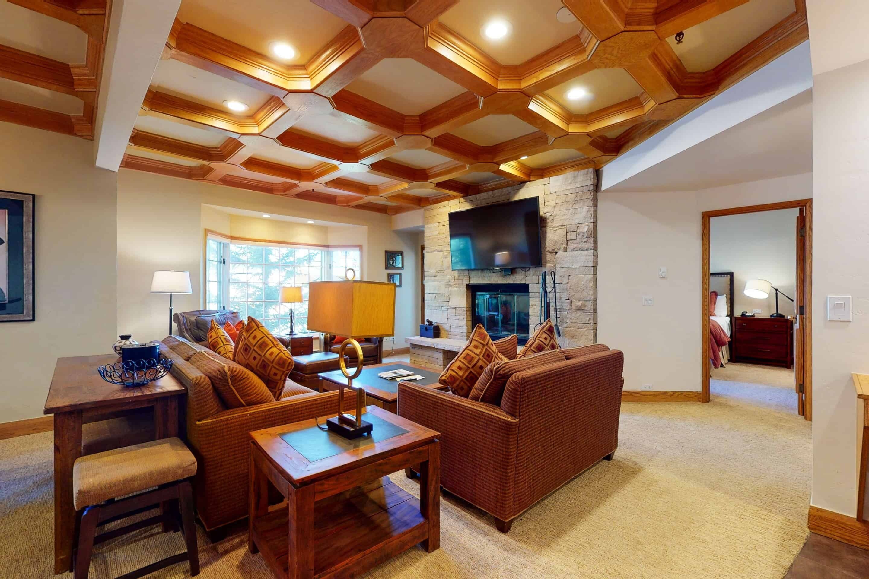 A spacious living room with a stone fireplace, wooden ceiling beams, and a large flat-screen TV mounted above it.