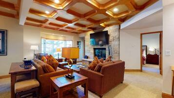 A spacious living room with a stone fireplace, wooden ceiling beams, and a large flat-screen TV mounted above it.