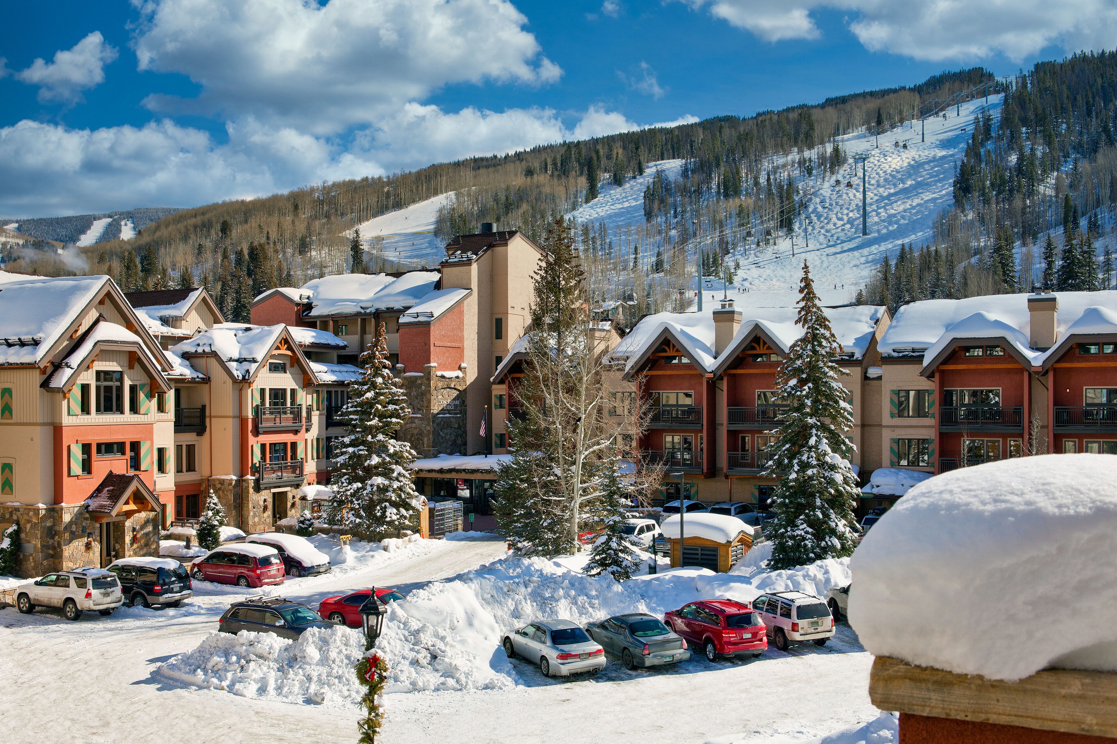 A snow-covered parking lot with several cars and a building with a ski slope in the background.