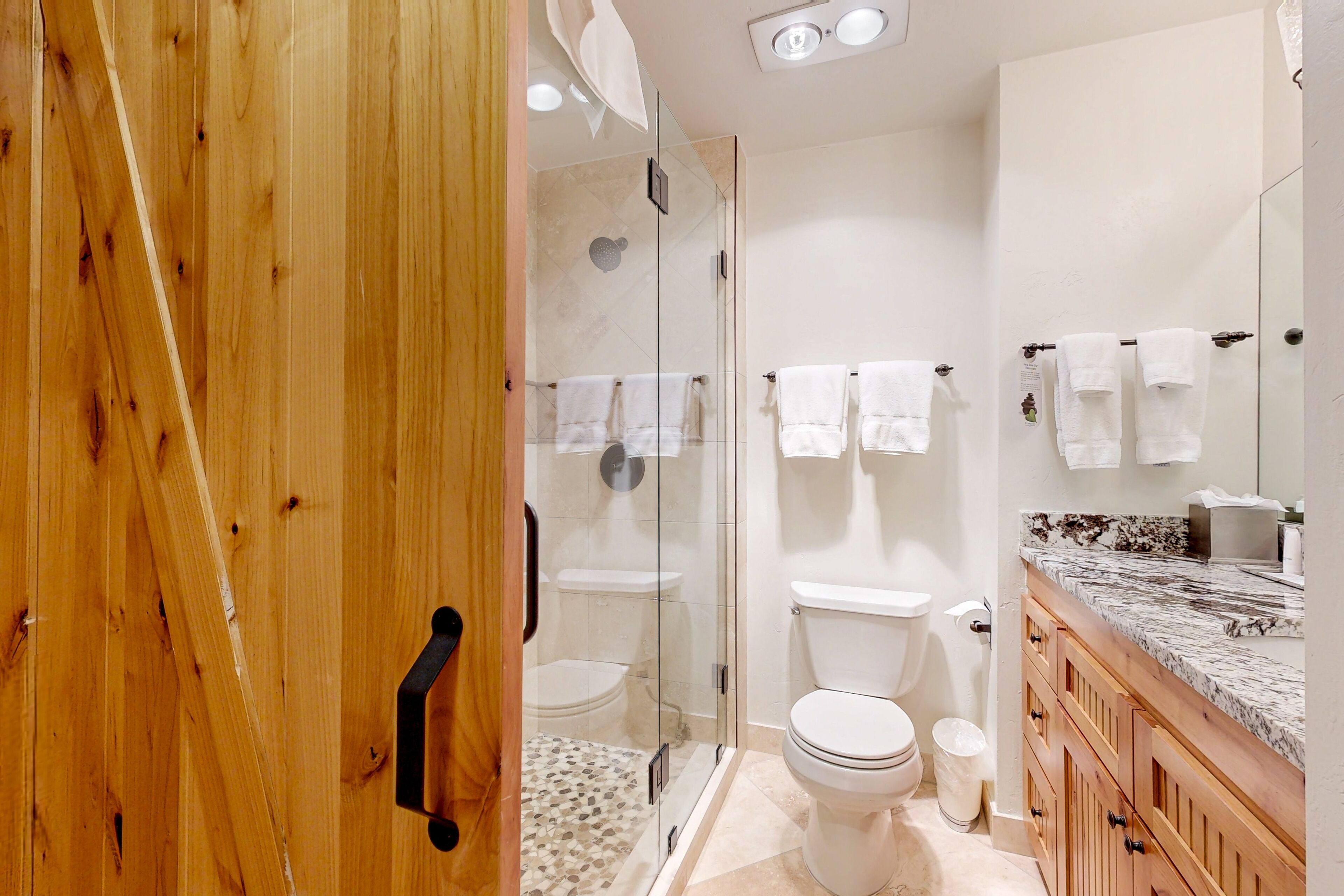 A modern bathroom with a glass shower enclosure, two toilets, a marble sink, and wooden cabinets.