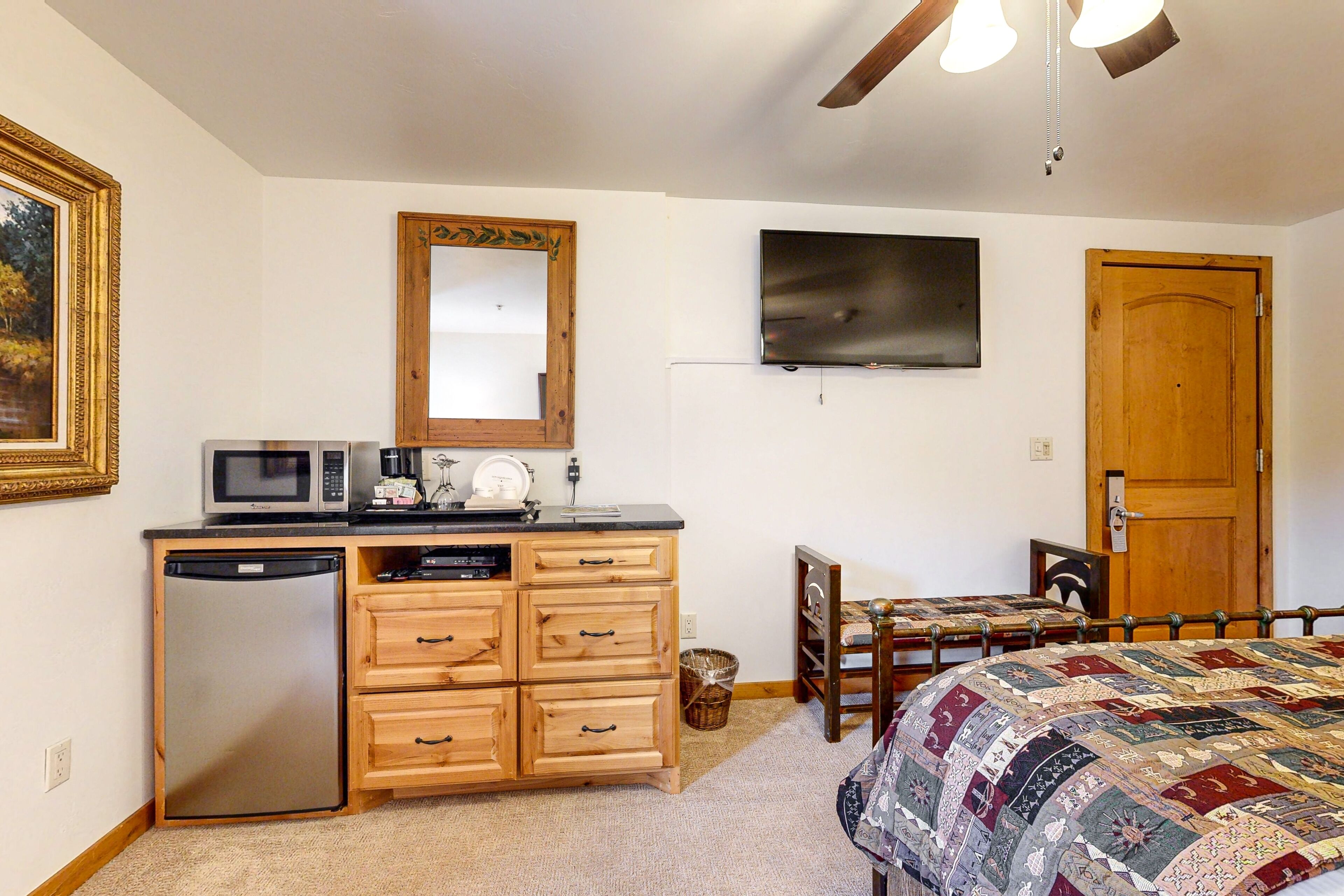 A bedroom with a bed, a wooden dresser, a microwave, a refrigerator, a television, and a wooden door.