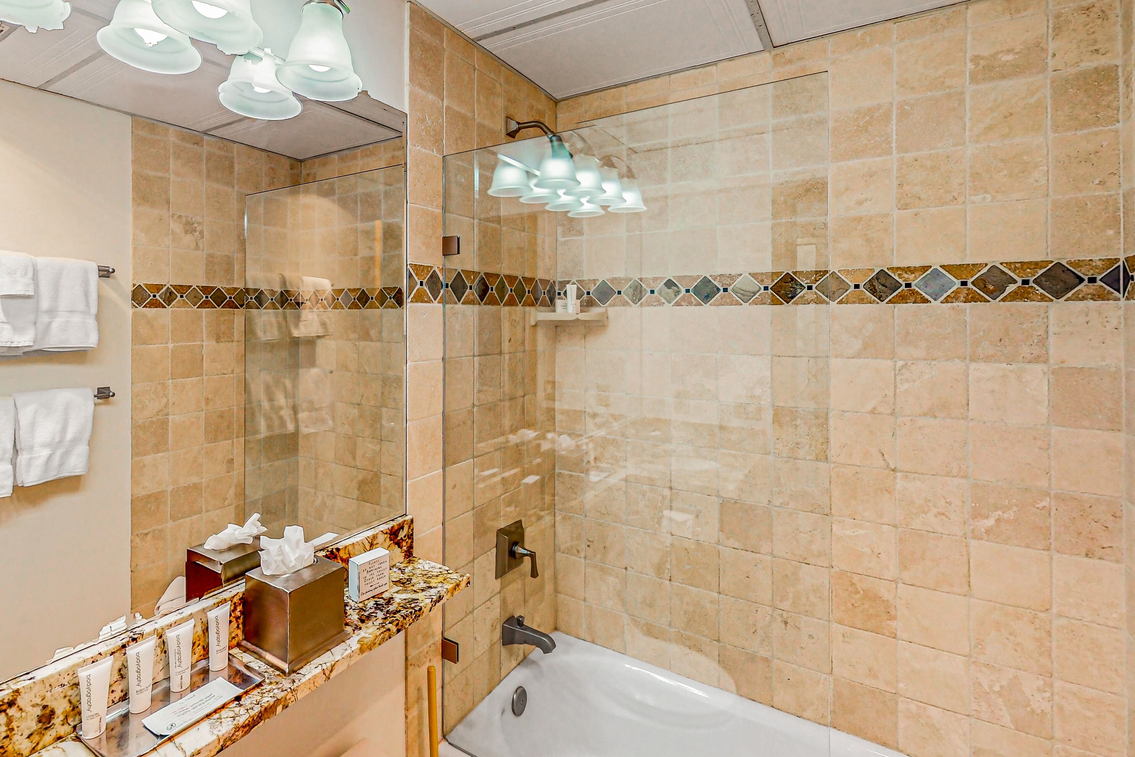 A bathroom with a marble countertop, a glass-enclosed shower, and a beige tiled wall.