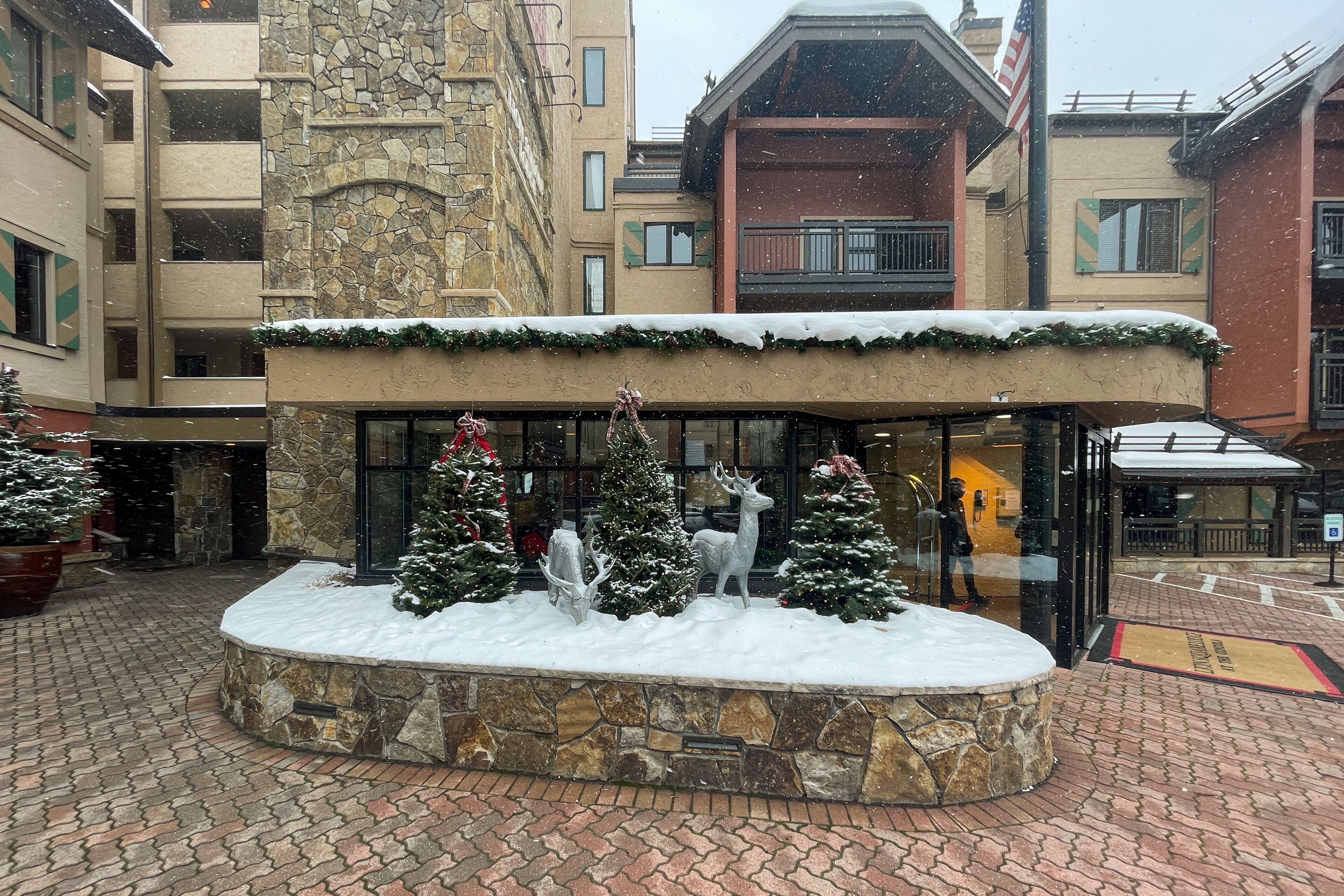 Un patio cubierto de nieve con una fuente de piedra, árboles de Navidad y la entrada de un edificio.