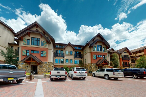 A multi-story hotel with a stone facade, multiple balconies, and a parking area with several vehicles.