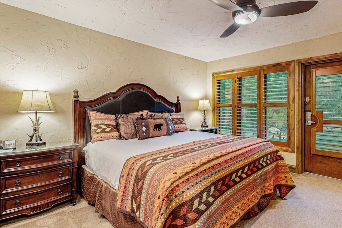 A bedroom with a large bed, a ceiling fan, a nightstand with a lamp, and a wooden door with shutters.