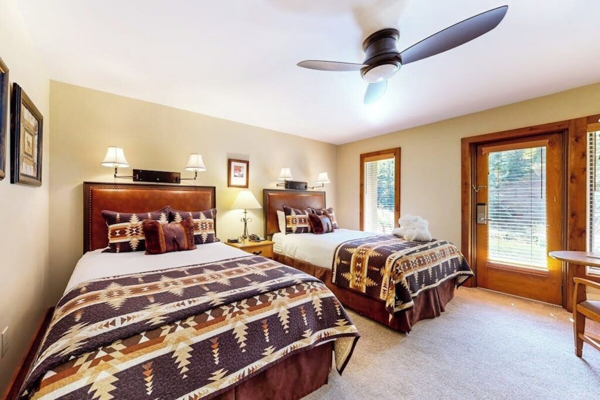 Habitación de hotel con dos camas, ventilador de techo, lámparas de noche, cabecero de madera, ventana con persianas y una pequeña mesa de madera.
