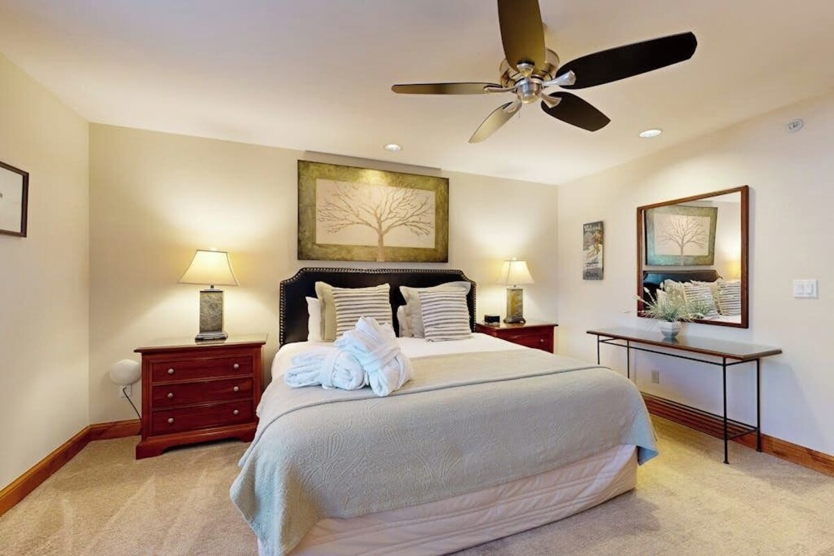 A hotel room with a bed, two bedside tables, a ceiling fan, a desk, and a chair.