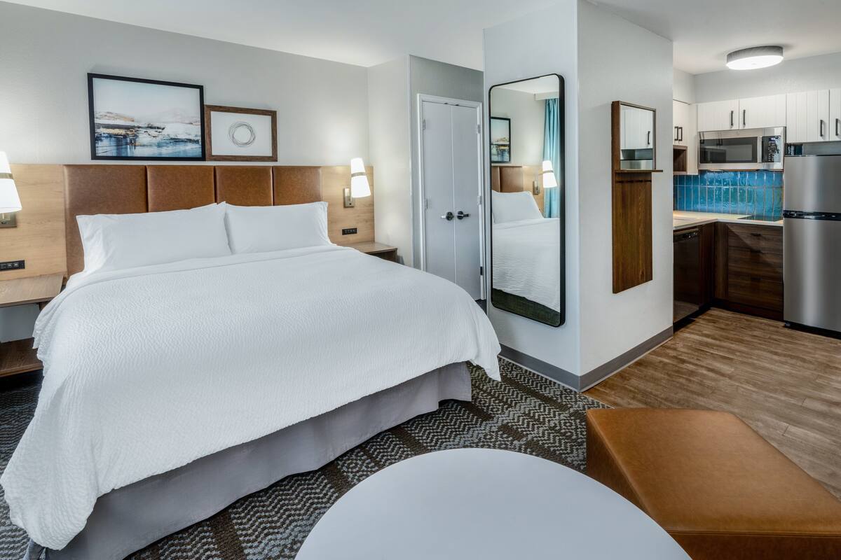 A hotel room with a large bed, a small desk, a mirror, a kitchenette, and a bathroom.
