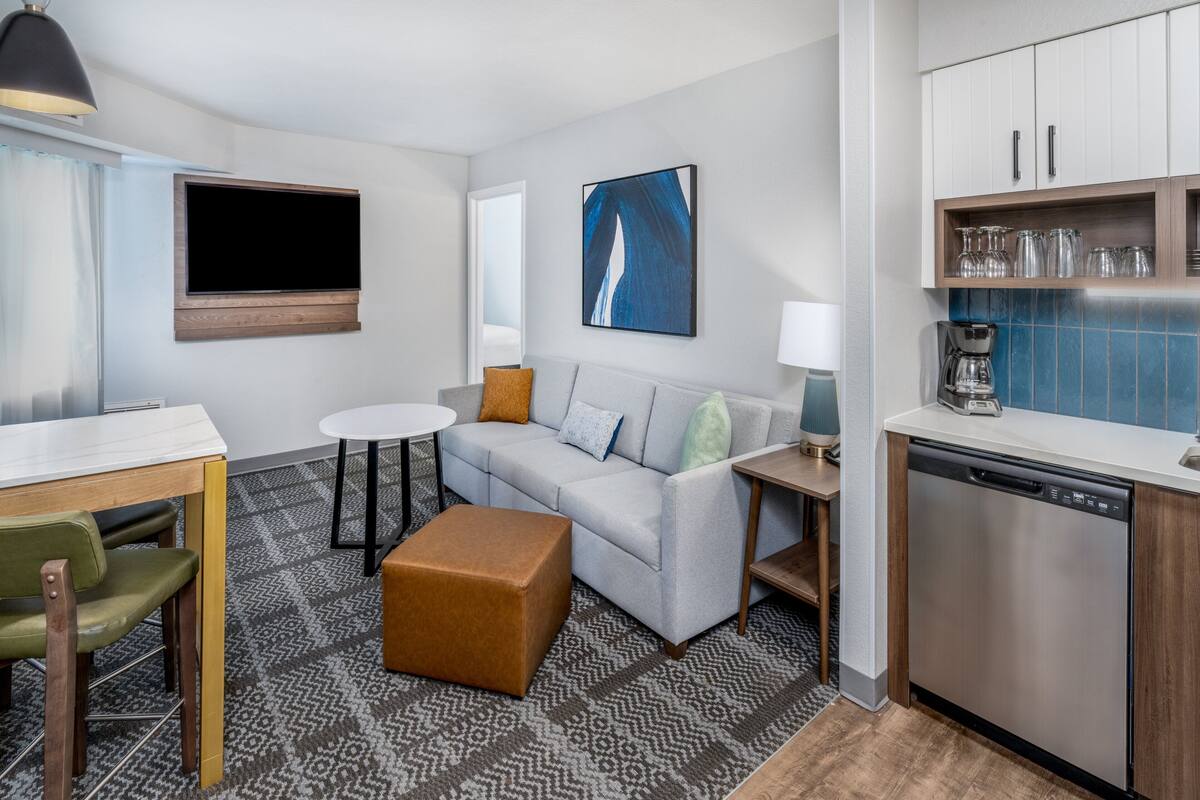 A modern hotel room with a sofa, a dining table, a coffee maker, and a dishwasher.