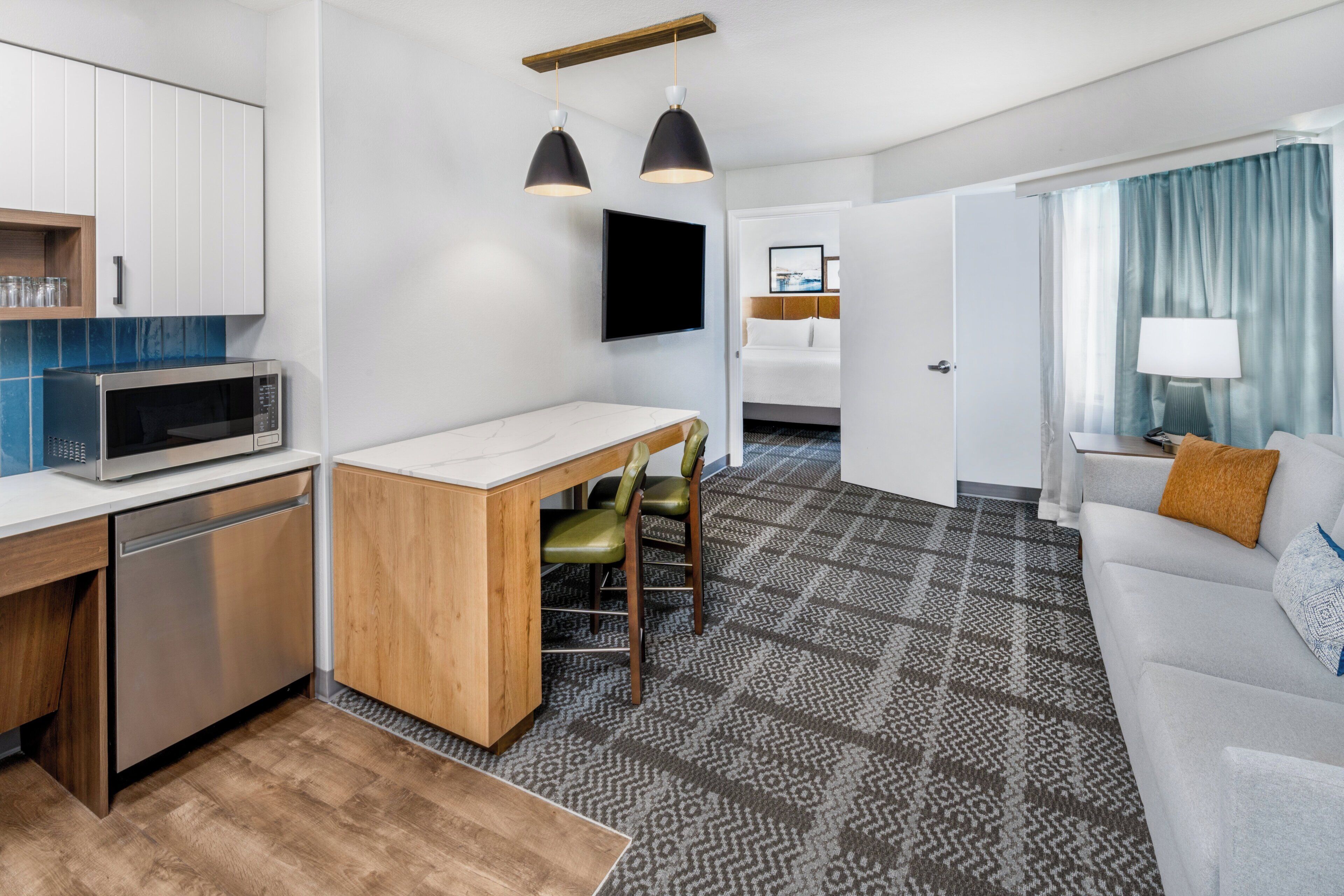 A compact hotel room with a kitchenette, a dining area, a living space with a sofa and a TV, and a patterned carpet.