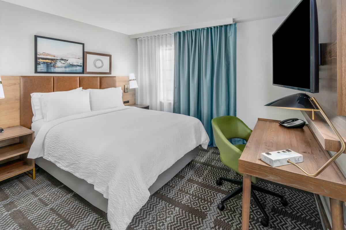 A hotel room with a large bed, a desk with a lamp, a chair, a television, and artwork on the wall.
