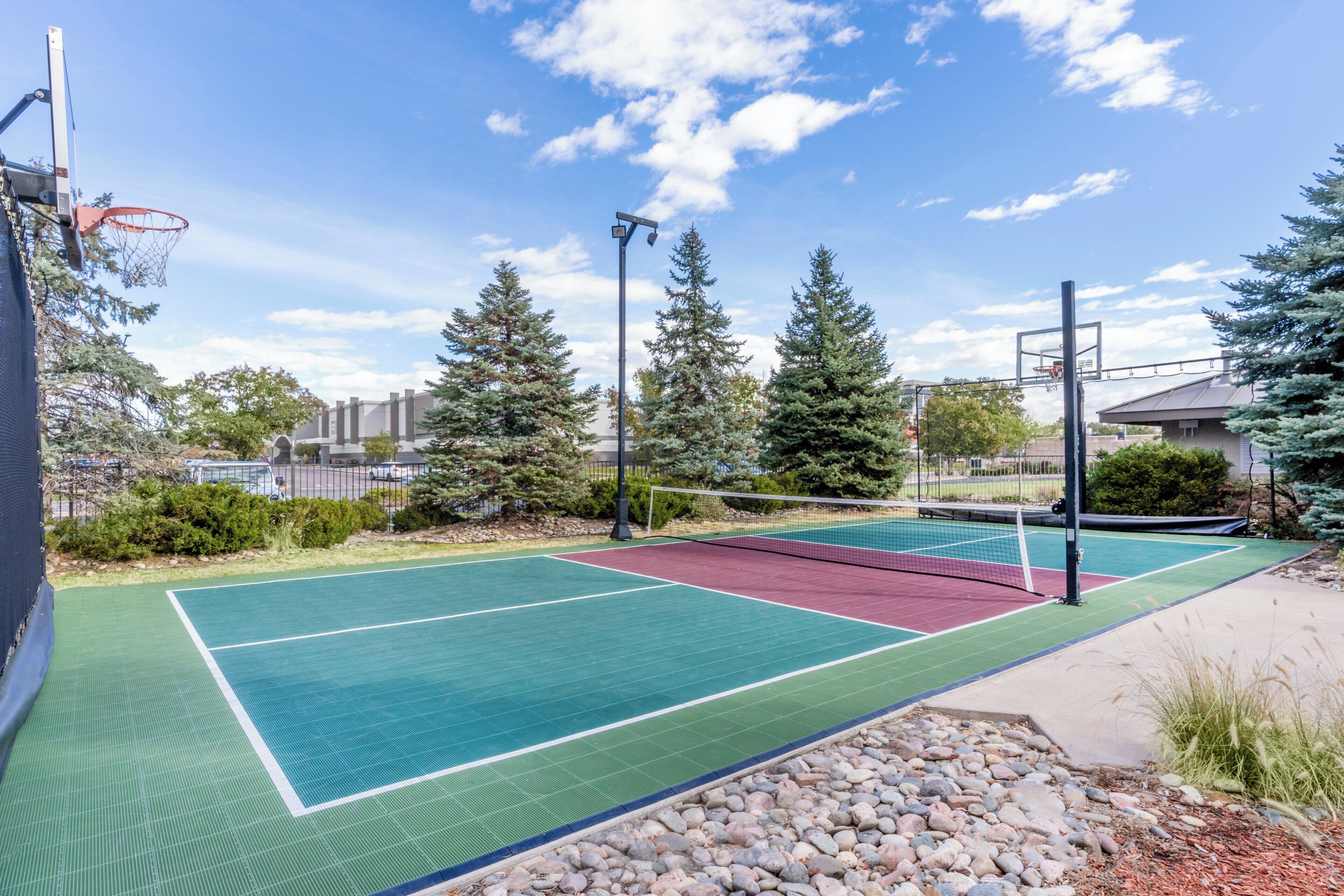 An outdoor tennis court with surrounding trees and a basketball hoop.