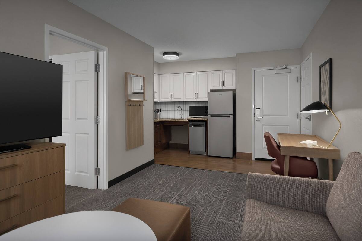 A modern hotel room with a flat-screen TV, a wooden cabinet, a sofa, a small table, and a kitchenette in the background.