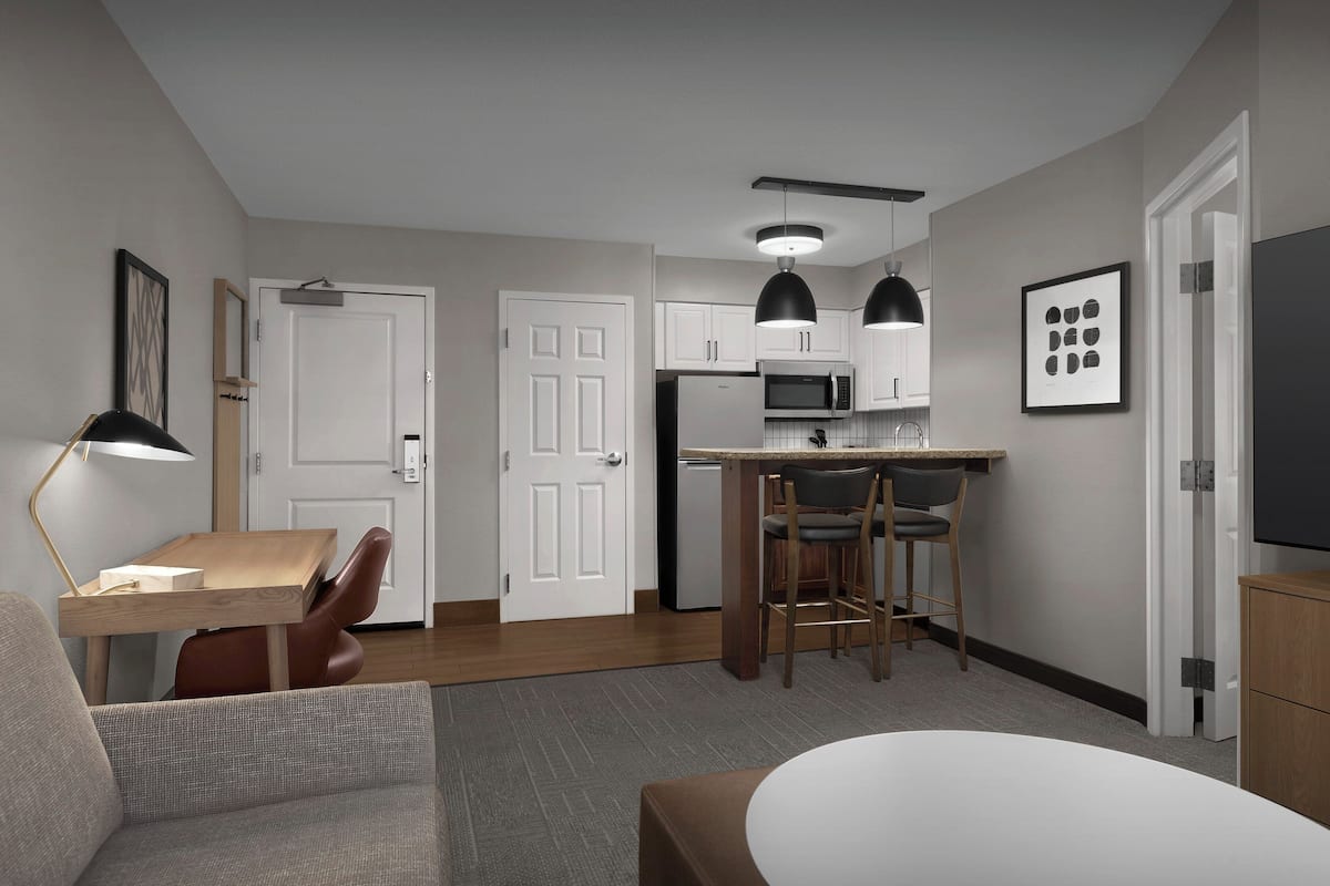 A modern kitchen with white cabinets, a microwave, and a bar stool. There is a dining table with chairs and a lamp.