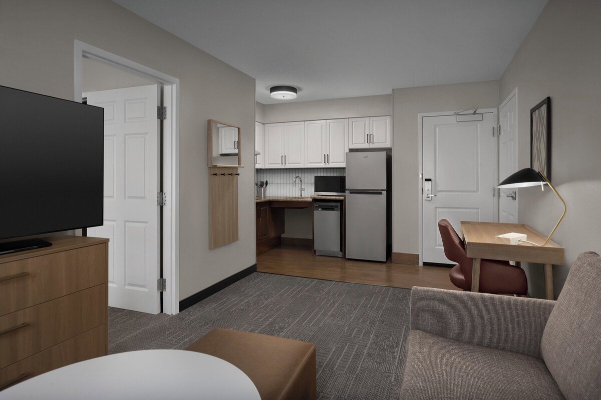 A modern hotel room with a flat-screen TV, a wooden cabinet, a sofa, a small table, and a kitchenette in the background.