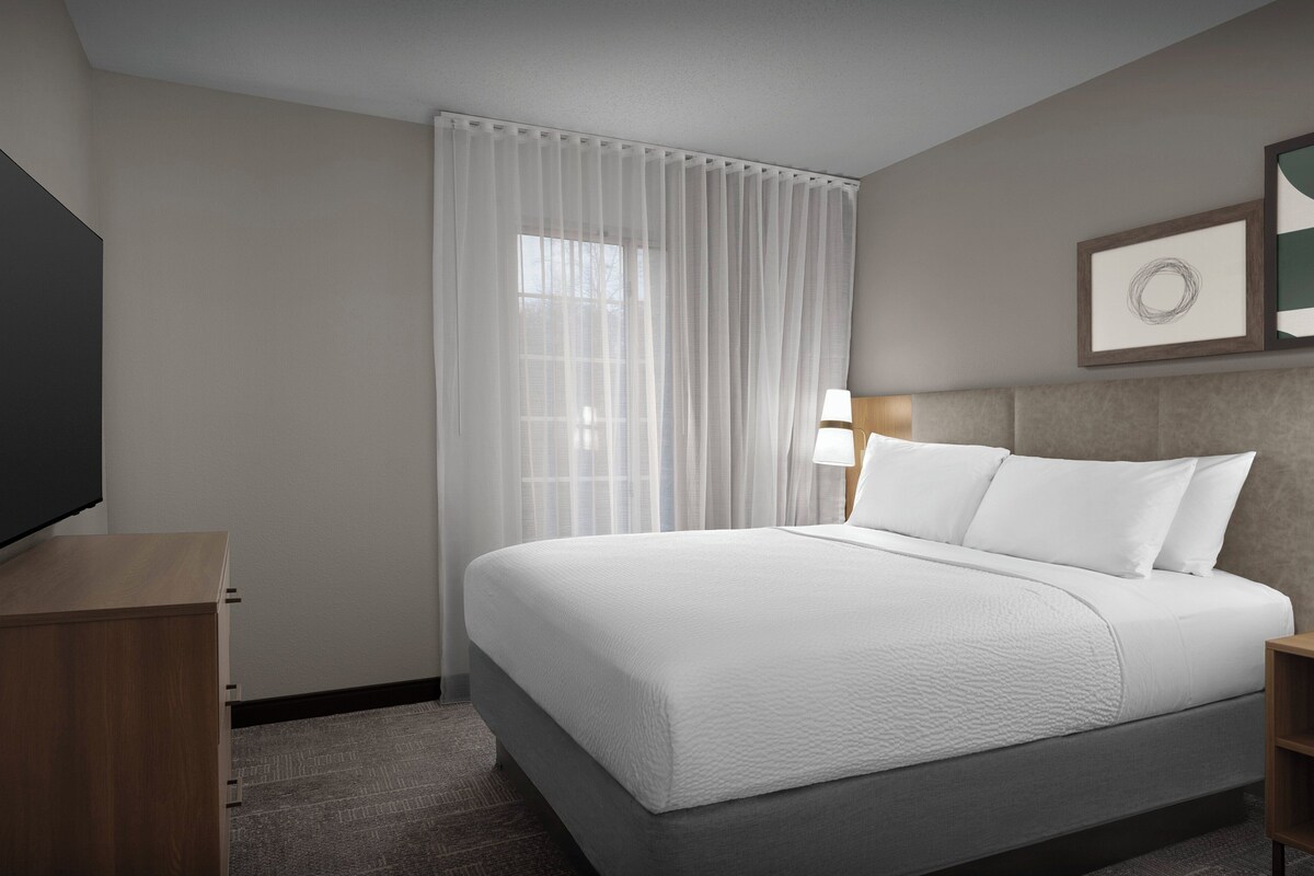 A hotel room with a large bed, a bedside table, a TV, and a window with sheer curtains.