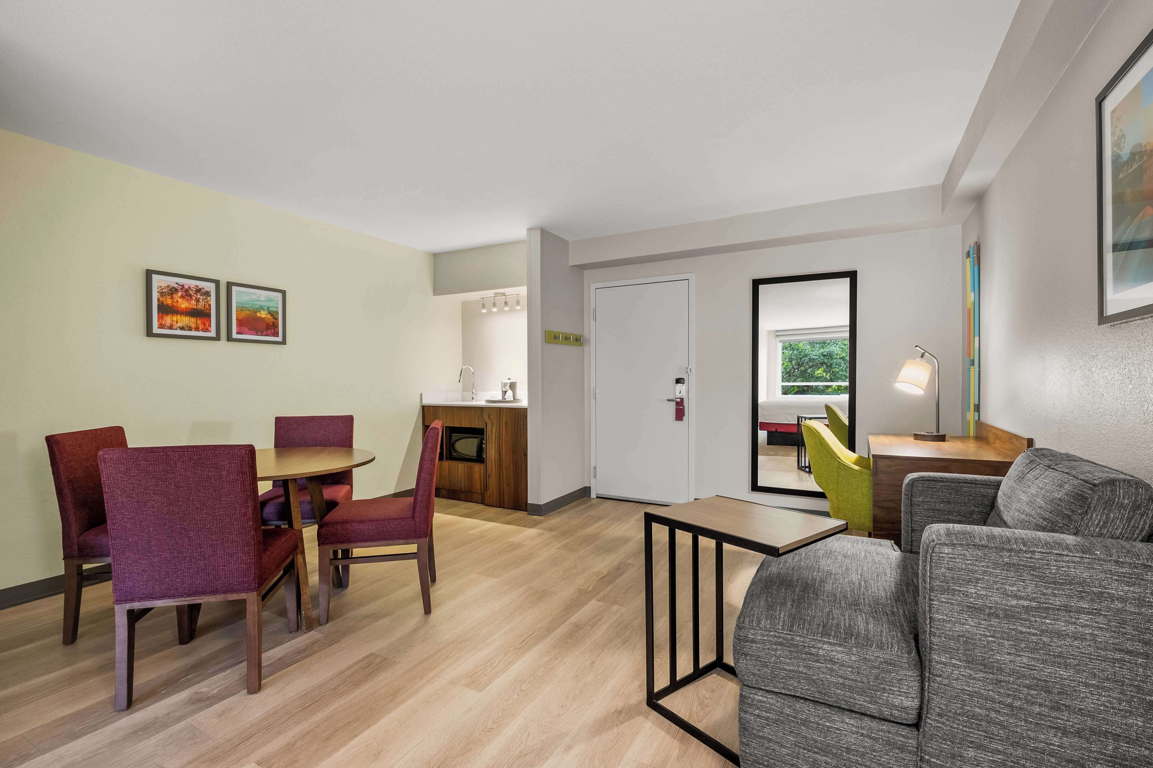 A modern living room with a grey sofa, a small table, and a dining area with red chairs. There is a kitchen area with a counter and a door leading to another room.
