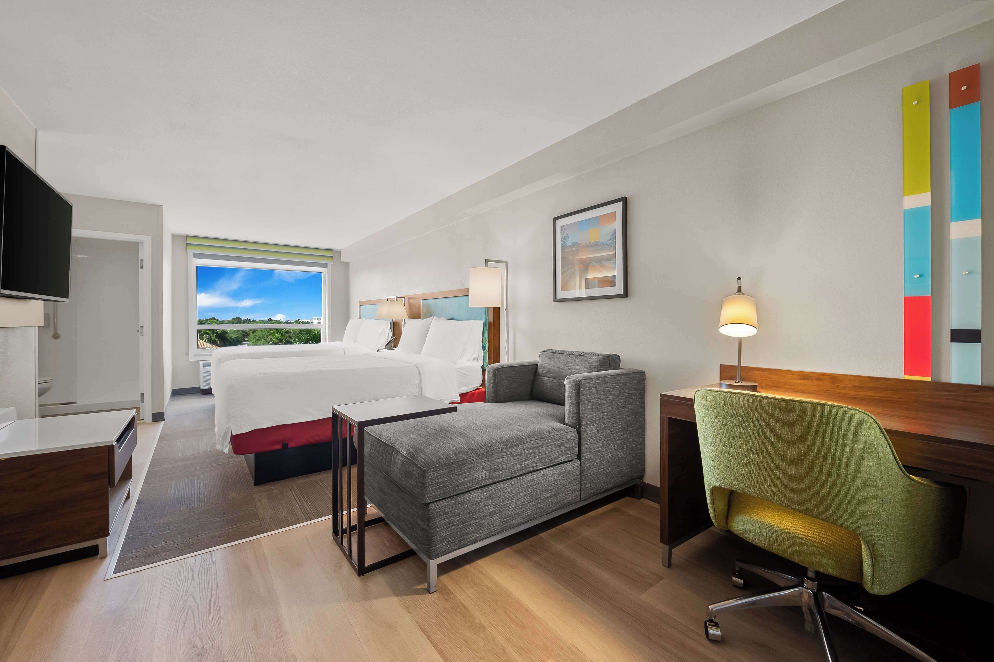 A hotel room with a bed, a desk, a chair, a TV, and a window with a view of greenery.