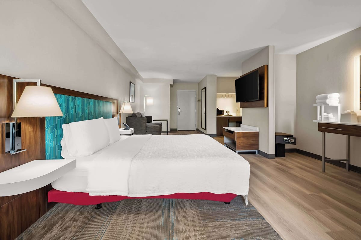 Una habitación de hotel moderna con cama, mesita de noche, lámpara, televisor y un pequeño escritorio con silla.