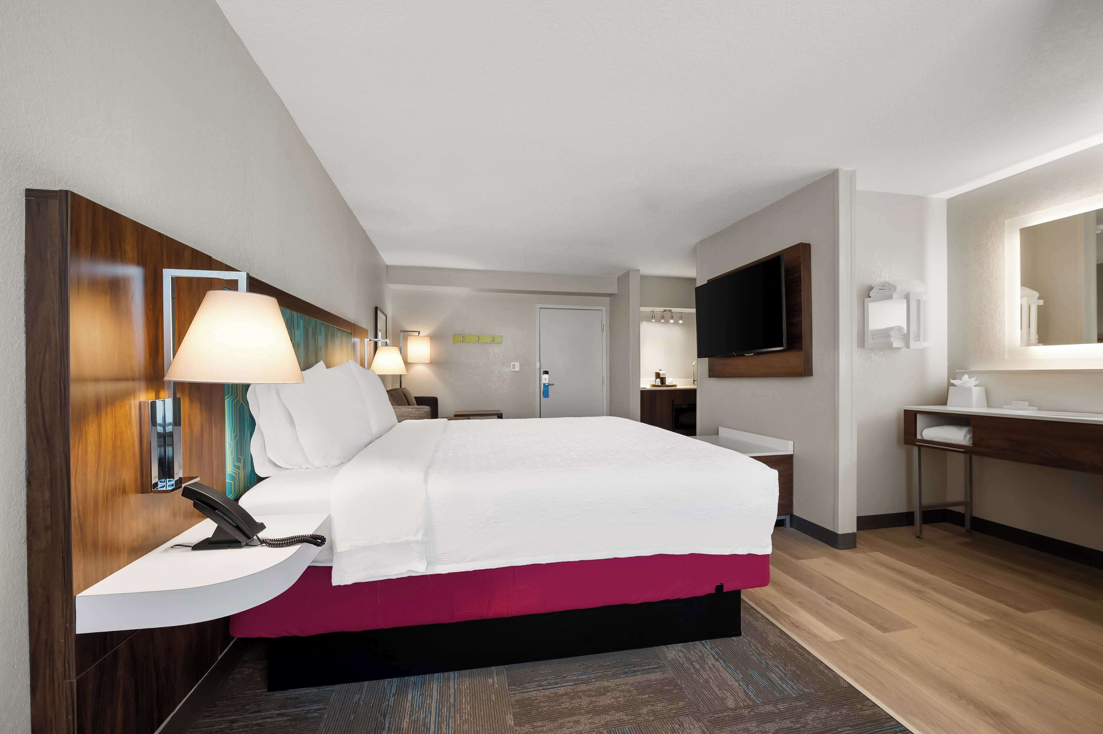 Une chambre d’hôtel comprenant un lit, une table de chevet, une lampe, une télévision et un bureau avec un miroir.