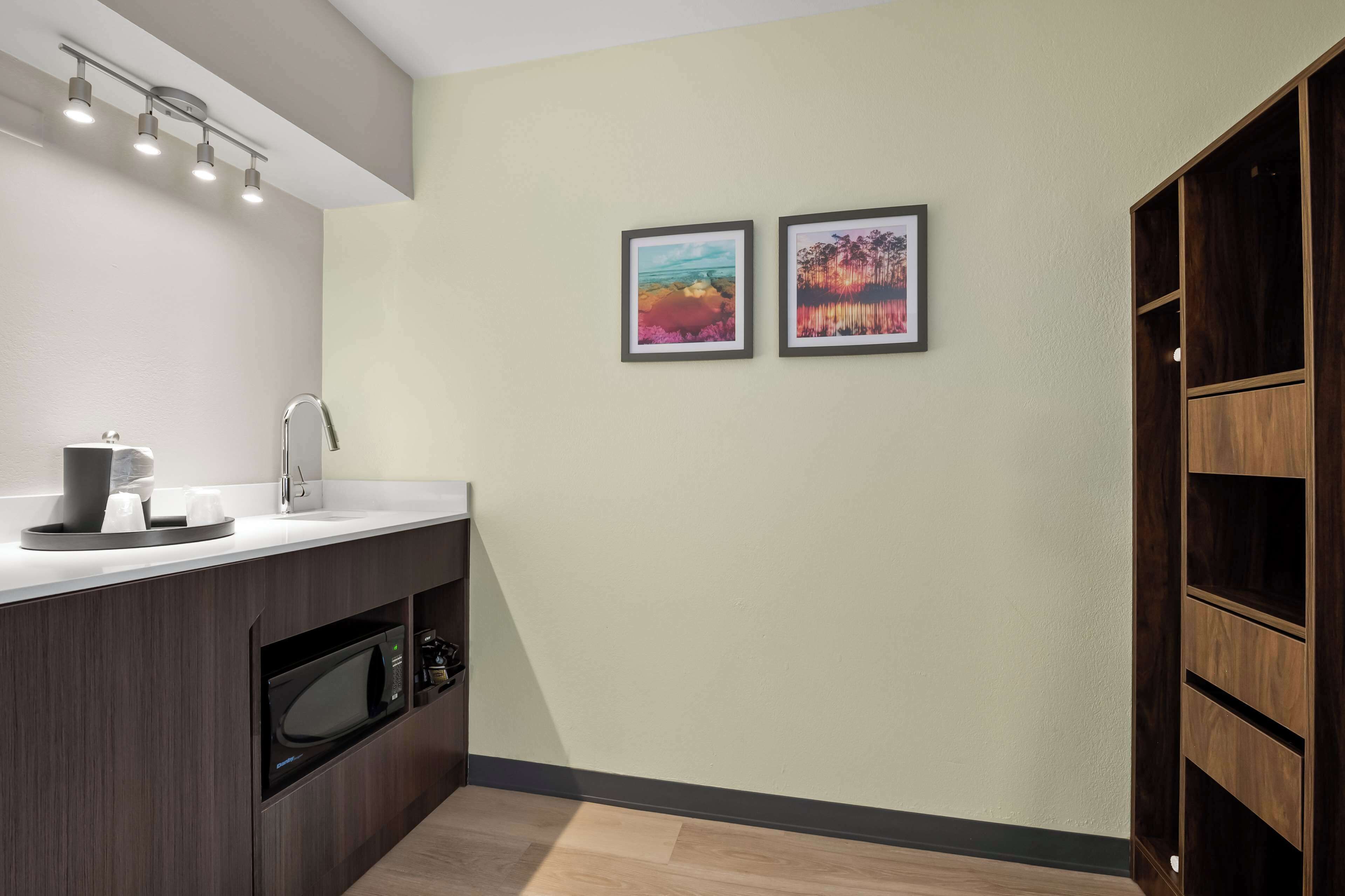 A modern kitchen with a built-in microwave, a sink, and a coffee maker. There are two framed pictures on the wall and a wooden cabinet to the right.