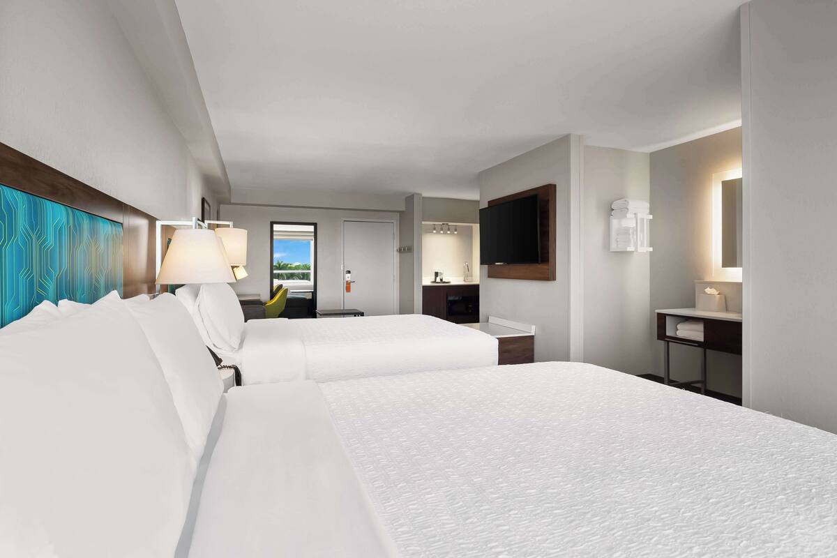Habitación de hotel con dos camas, un escritorio, un televisor y un baño visible a través de una puerta abierta.