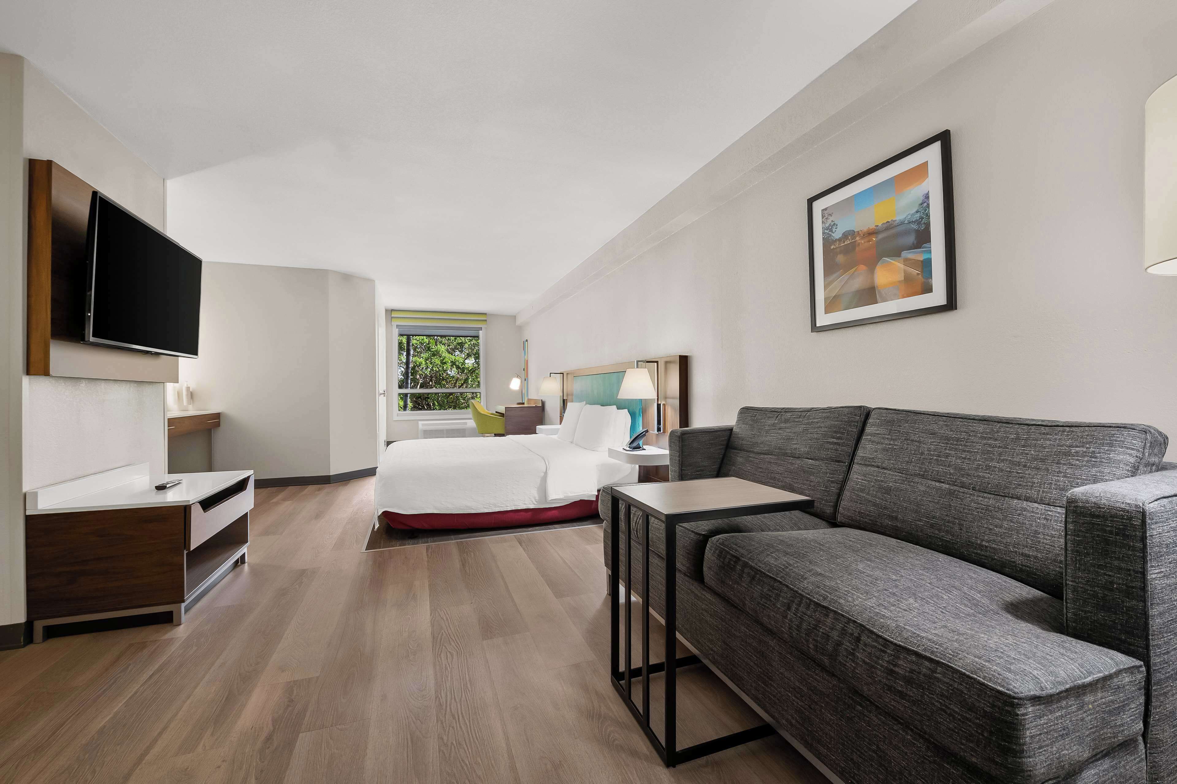 Et moderne hotelværelse med et fladskærms-tv, en grå sofa, en seng med hvide sengetøj, et lille bord og et vindue med udsigt over grønne områder.
